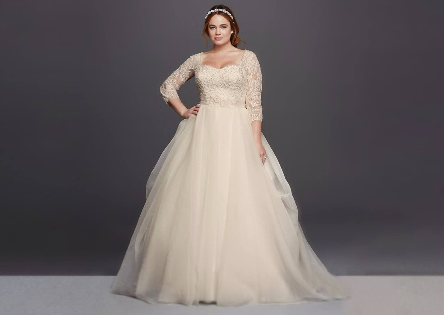 Koyu renk arka plan önünde duran gelinin üzerinde kabarık etekli bir gelinlik bulunuyor.