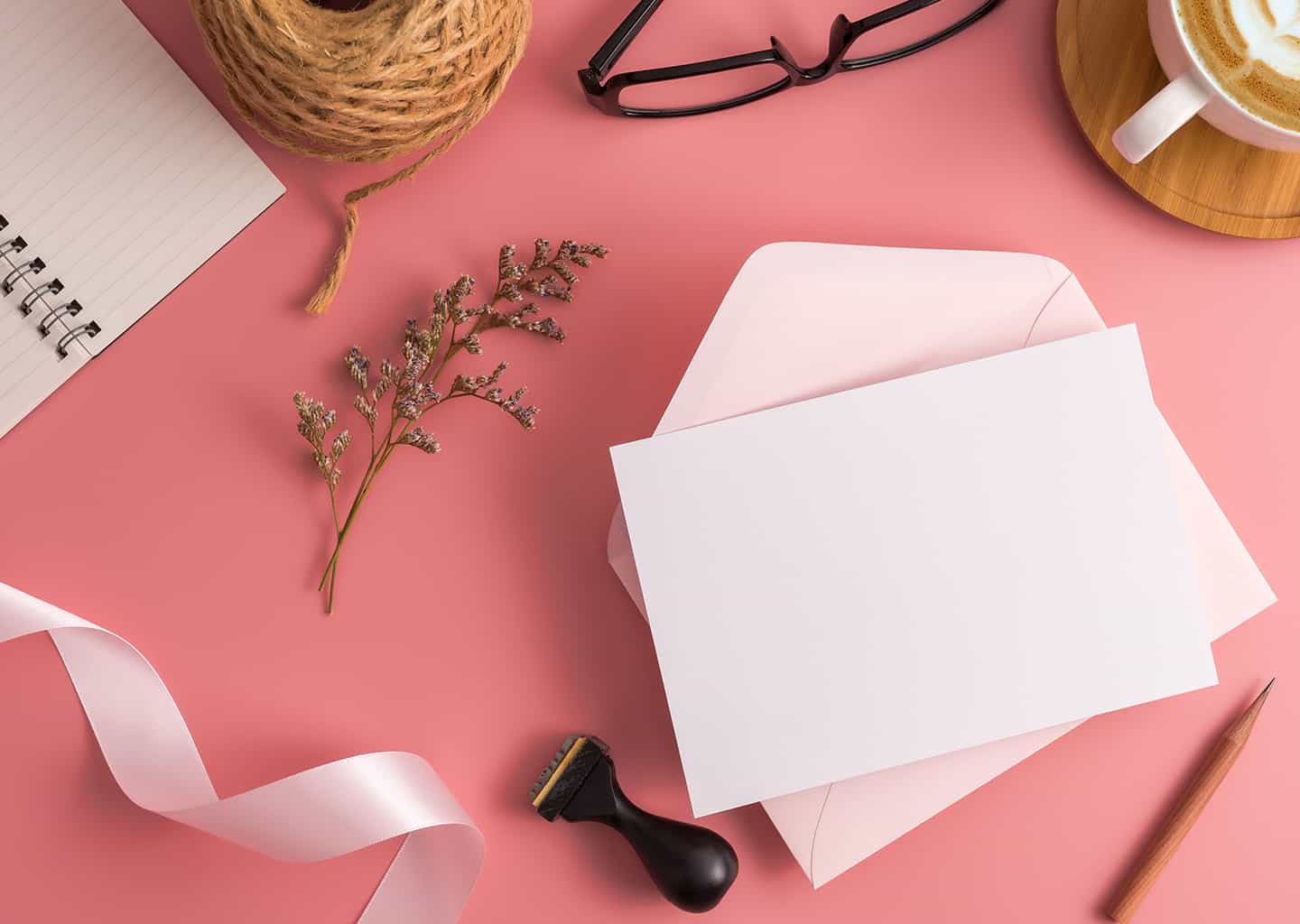 Koyu pembe arka plan üzerinde bol bir not defteri, boş bir davetiye, pembe bir kurdele, kurşun kalem, gözlük, ip, içi kahve ile dolu bir fincan ve damga bulunuyor.
