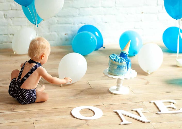 Ahşap zemin üzerinde arkası dönük bir bebek oturuyor. Zemin üzerinde beyaz, mavi renkte balonlar ve mavi renkte bir pasta bulunuyor.