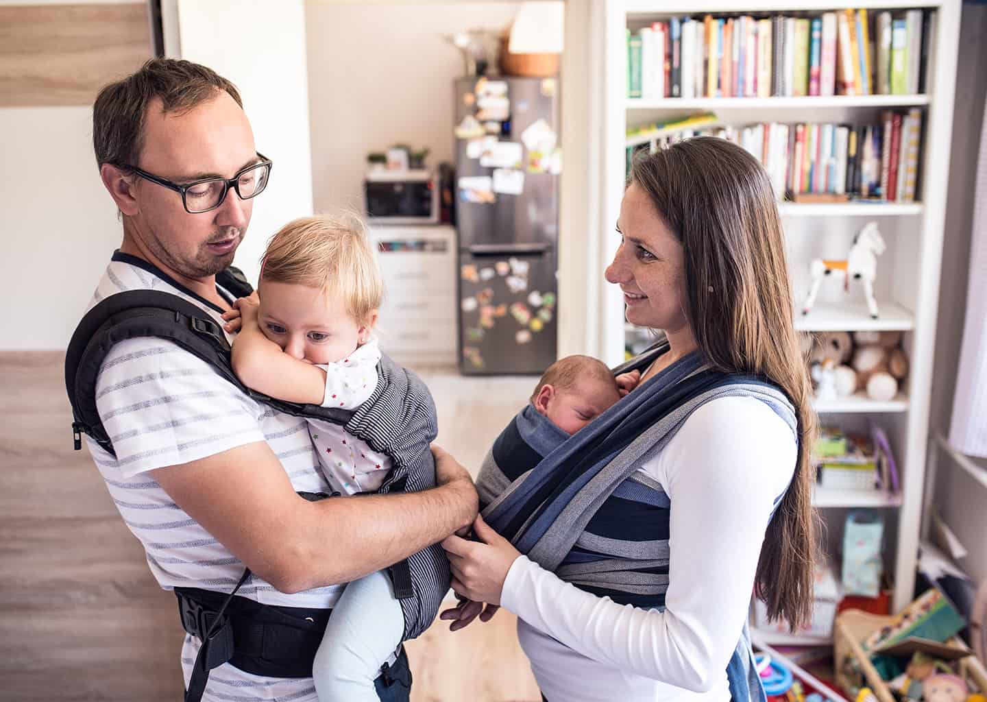 Oturma odasında duran genç kadın ve adam kucaklarında biri bebek olan çocuklarını tutuyorlar. 