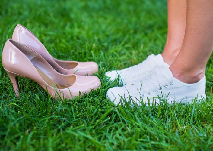 Yakın plan çekimde çimlerin üzerinde bir çift topuklu ayakkabı duruyor. Topuklu ayakkabıların karşısında ise spor ayakkabı giymiş bir kadın duruyor.