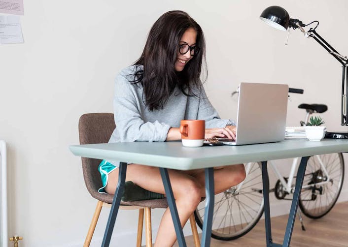 Genç kadın çalışma masasında otururken, masa üzerinde bulunan dizüstü bilgisayarda yazı yazıyor. Masada bir kupa, çiçek ve lamba bulunuyor.
