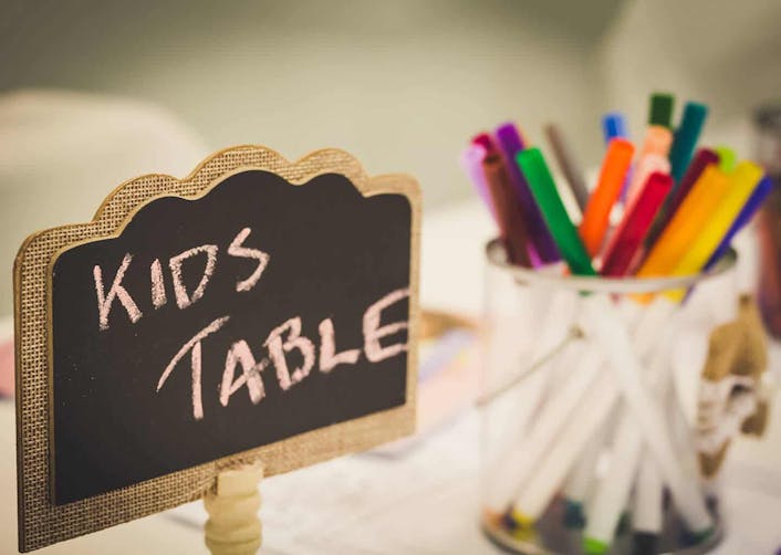 Yakın plan çekimde; küçük siyah renkli bir tahta üzerinde "Kids Table" yani "Çocukların Masası" yazıyor. Tahtanın yanında, içinde renkli boya kalemlerinin bulunduğu şeffaf bir kalemlik duruyor.