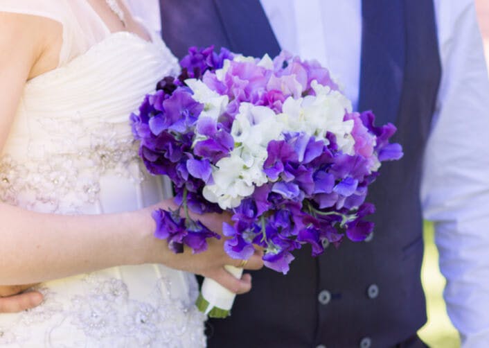 Yakın plan çekimde gelin ve damat birbirine sarılıyor. Gelin elinde mor ve beyaz renkli bezelye çiçeğinden yapılmış bir buket tutuyor.