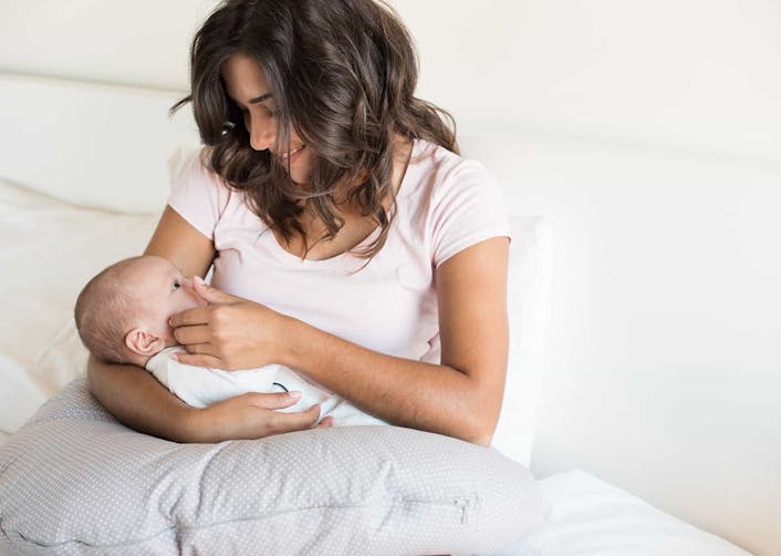 Genç kadın yatağında otururken, kolları ile sardığı bebeğini seviyor ve gülümsüyor. Bebeğin altında bir yastık bulunuyor.