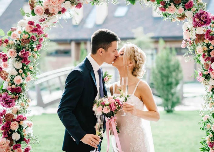 Düğün salonunun bahçesinde, çiçeklerden taç şeklinde yapılmış dekor altında duran gelin ve damat, birbirini öpüyor.