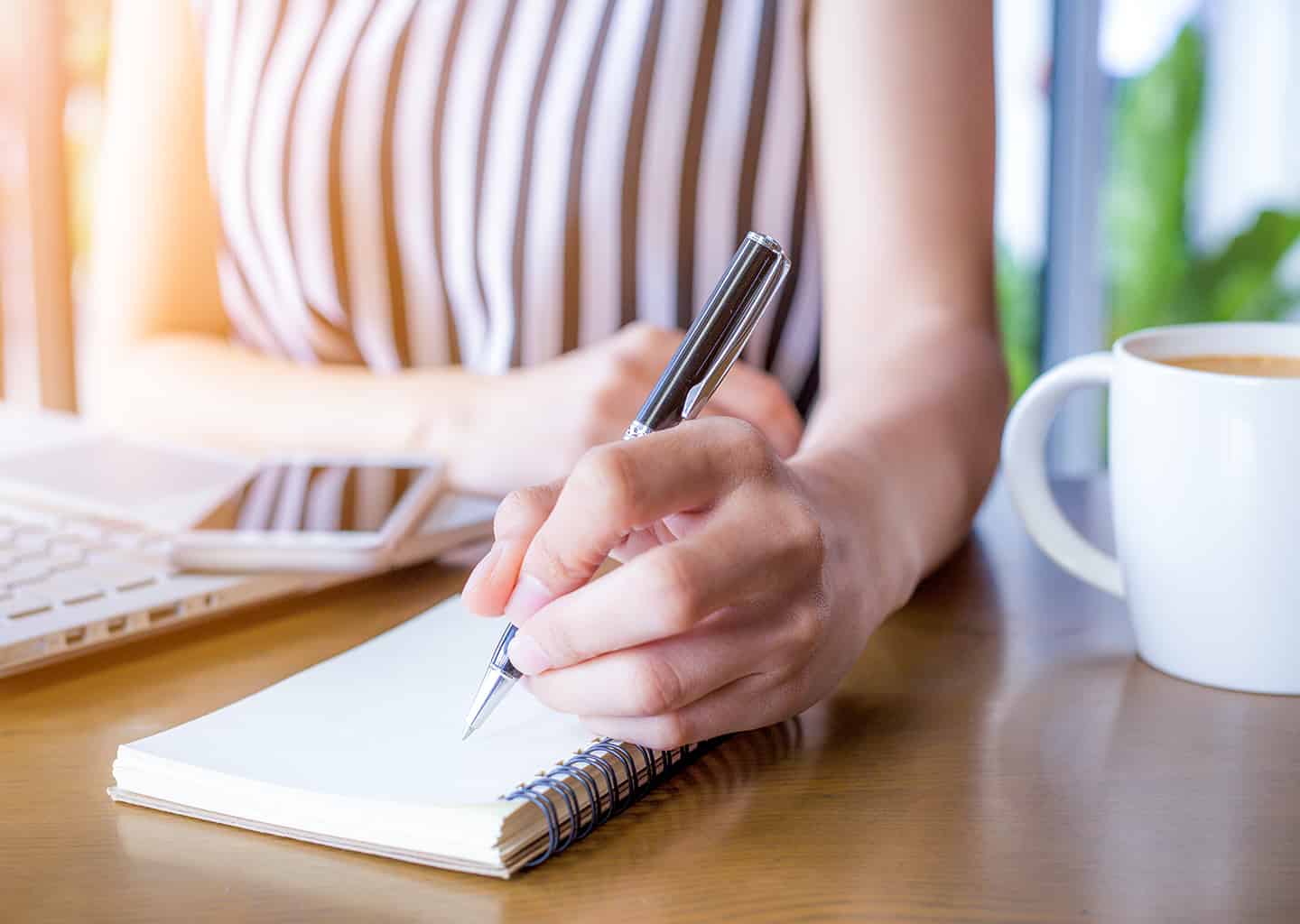 Yakın plan çekimde kadın masasında otururken not defterine notlar alıyor. Masada bir laptop ve bir kupa içerisinde kahve bulunuyor.