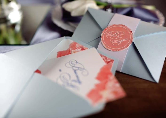 Yakın plan çekimde açık mavi renkte iki adet düğün davetiye zarfı bulunuyor. Zarflardan birinin içerisinden beyaz ve pembe renkler ile hazırlanmış düğün davetiyesi görünüyor.