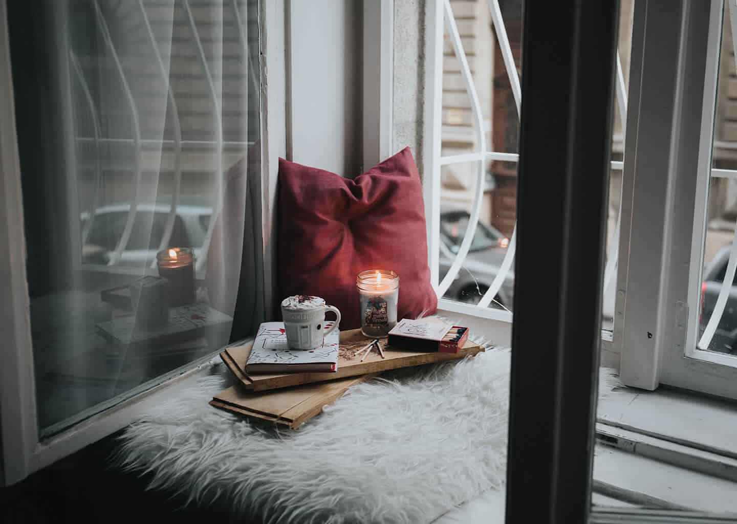 Pencerenin önünde kırmızı bir yastık, yanan bir mum, kulplu bir fincan, açık renk peluş ve tahtalar bulunuyor.