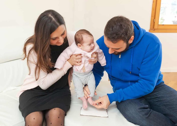 Açık renk koltuk üzerinde bir kadın ve adam oturuyor. Kadın iki eli ile birlikte bebeğini tutarken, adam ise bebeğinin ayaklarını tutarak bebeğinin ayak kalıbını alıyor.