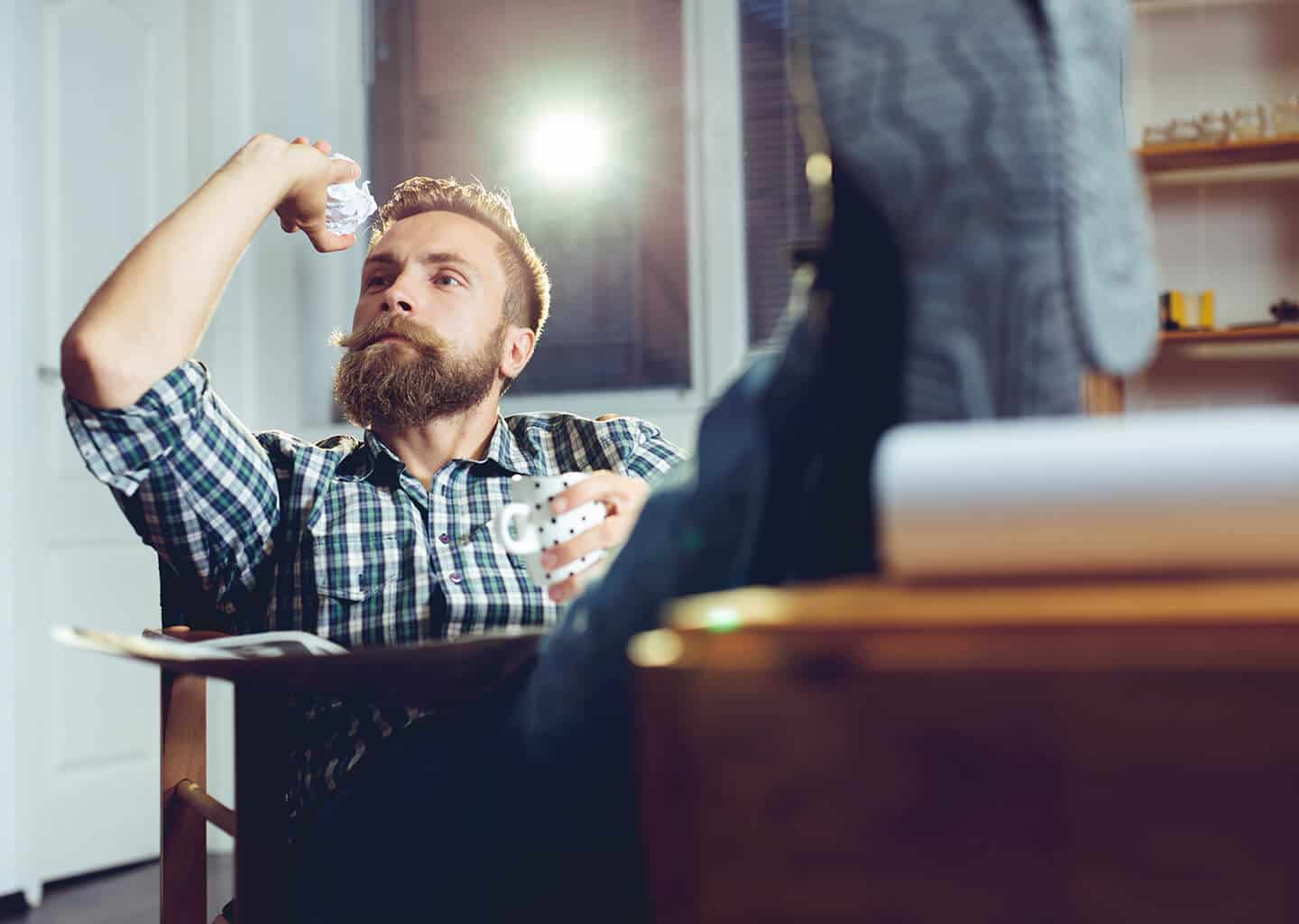 Tahta bir sandalyede oturan ve ayağını masaya uzatmış olan genç adam, elinde buruşturmuş olduğu kağıt parçasını fırlatmak için kolunu kaldırıyor.