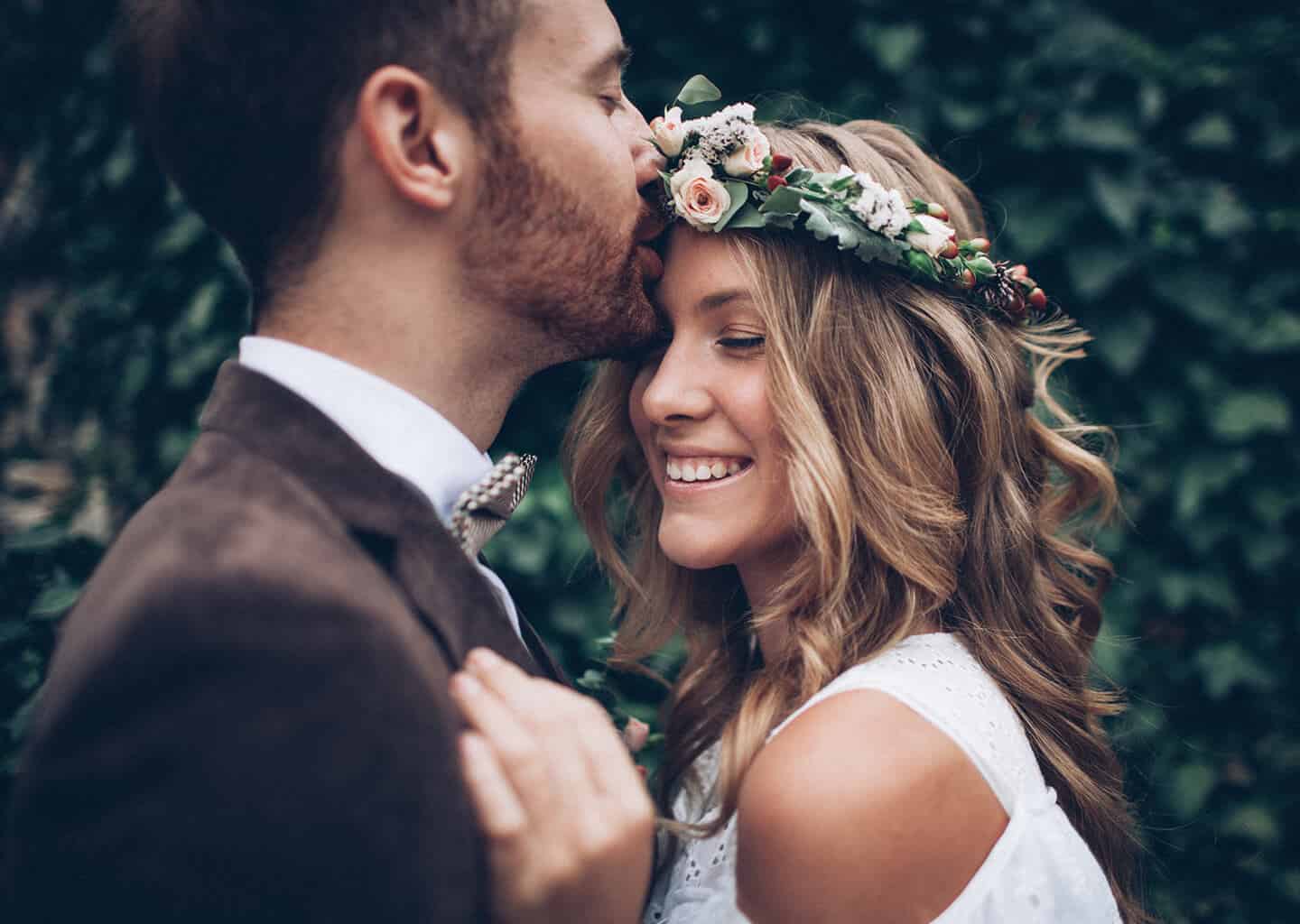 Yakın plan çekimde, yeşillik bir arka planın önünde damat gelini alnından öpüyor, gelin gözleri kapalı bir şekilde gülümsüyor. Gelinin başında çiçeklerden yapılmış bir taç bulunuyor.
