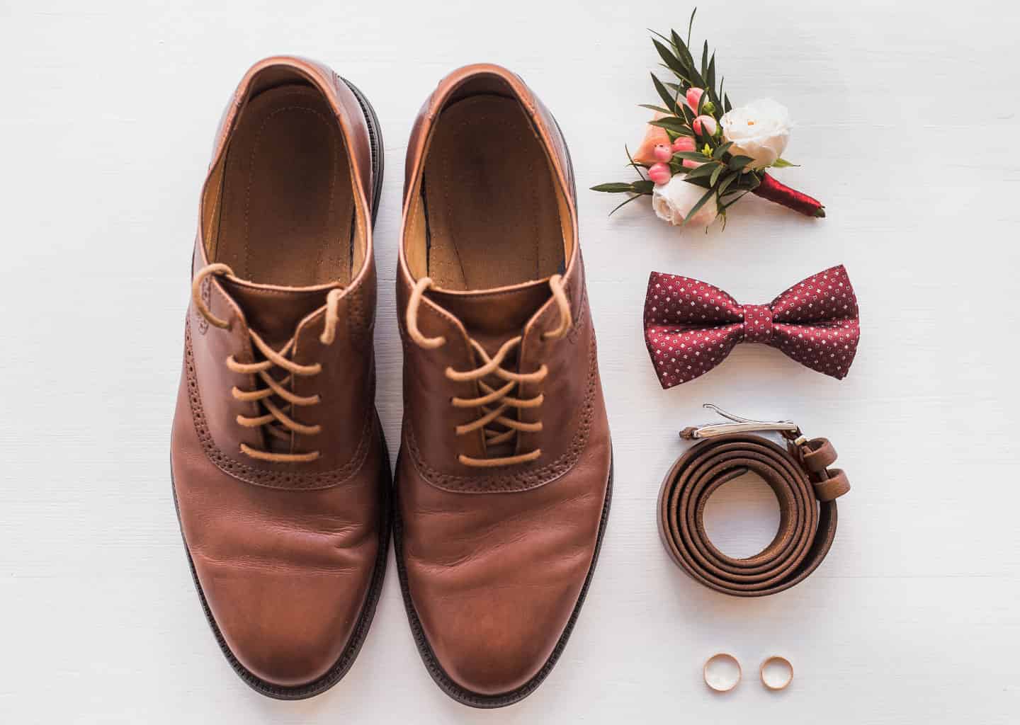 Açık renk arka plan üzerinde kahverengi bir erkek ayakkabısı, kırmızı bir papyon, kahverengi kemer, alyanslar ve cep çiçeği bulunuyor.