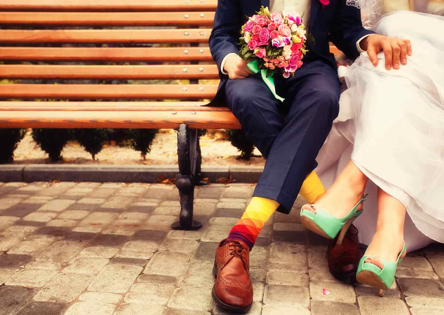 Yakın plan çekimde gelin ve damat bir bank üzerinde oturuyor. Damadın elinde gelin çiçeği bulunuyor.