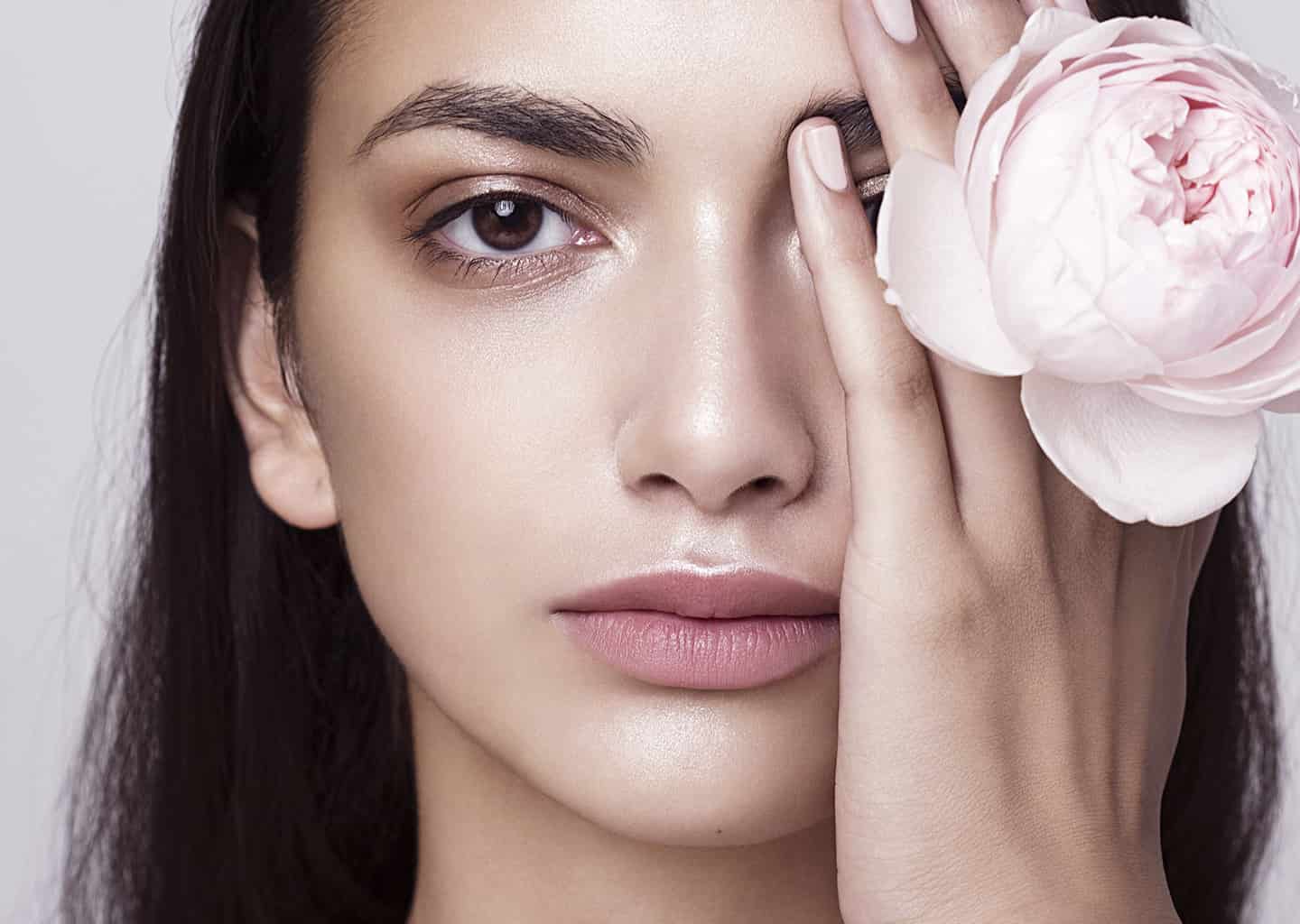 Yakın plan çekimde genç kadın sol eli ile sol gözünü kapatıyor. Kadının sol elinde, parmakları arasında sıkıştırılmış açık pembe bir gül bulunuyor.