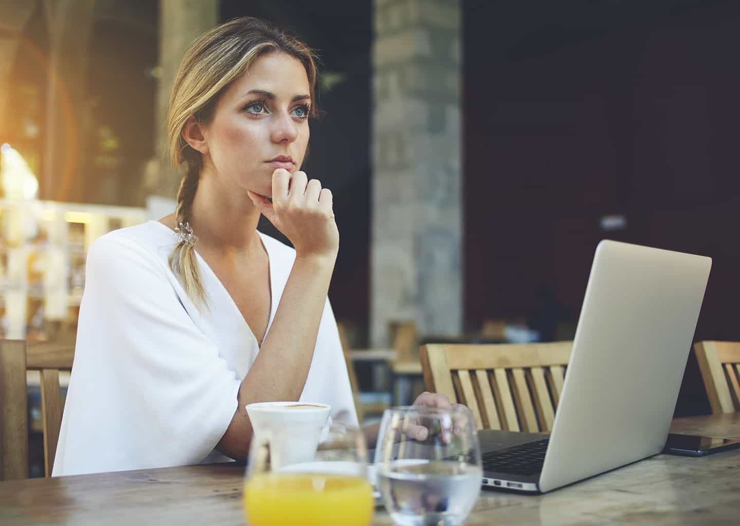 Genç kadın yemek masasında otururken sağ eli ile çenesini tutarak ve karşıya bakarak düşünüyor. Masanın üzerinde dizüstü bilgisayar, su bardağı ve limonata bardağı bulunuyor.