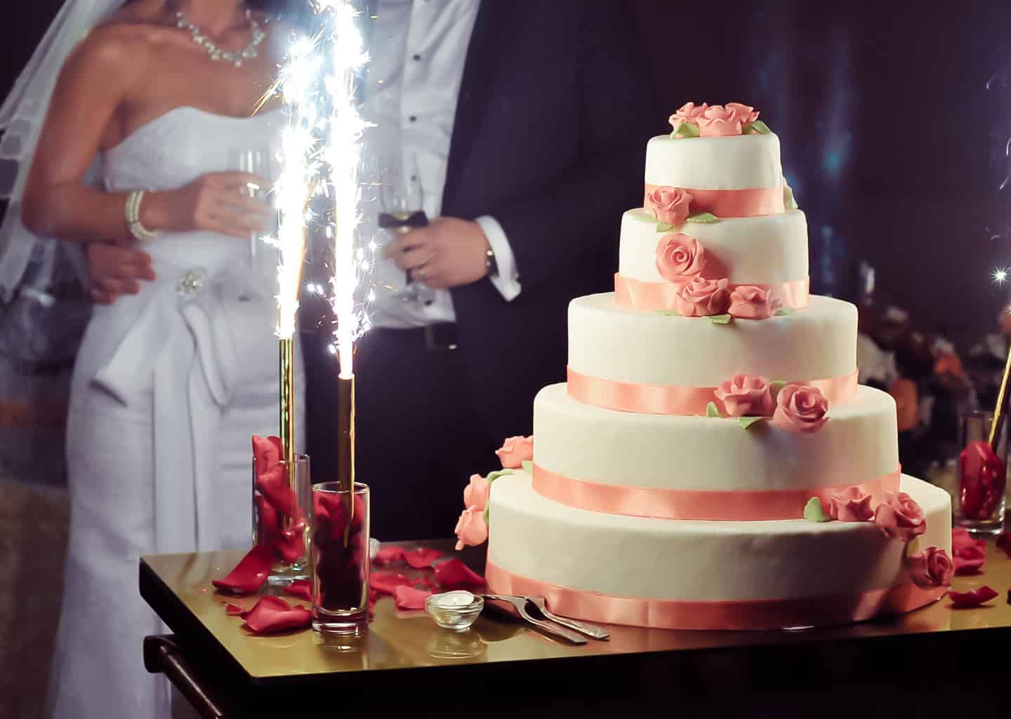 Yakın plan çekimde gelin ve damat; beş katlı olan düğün pastalarına bakıyor. Gelin ve damadın elinde kadeh bulunuyor.