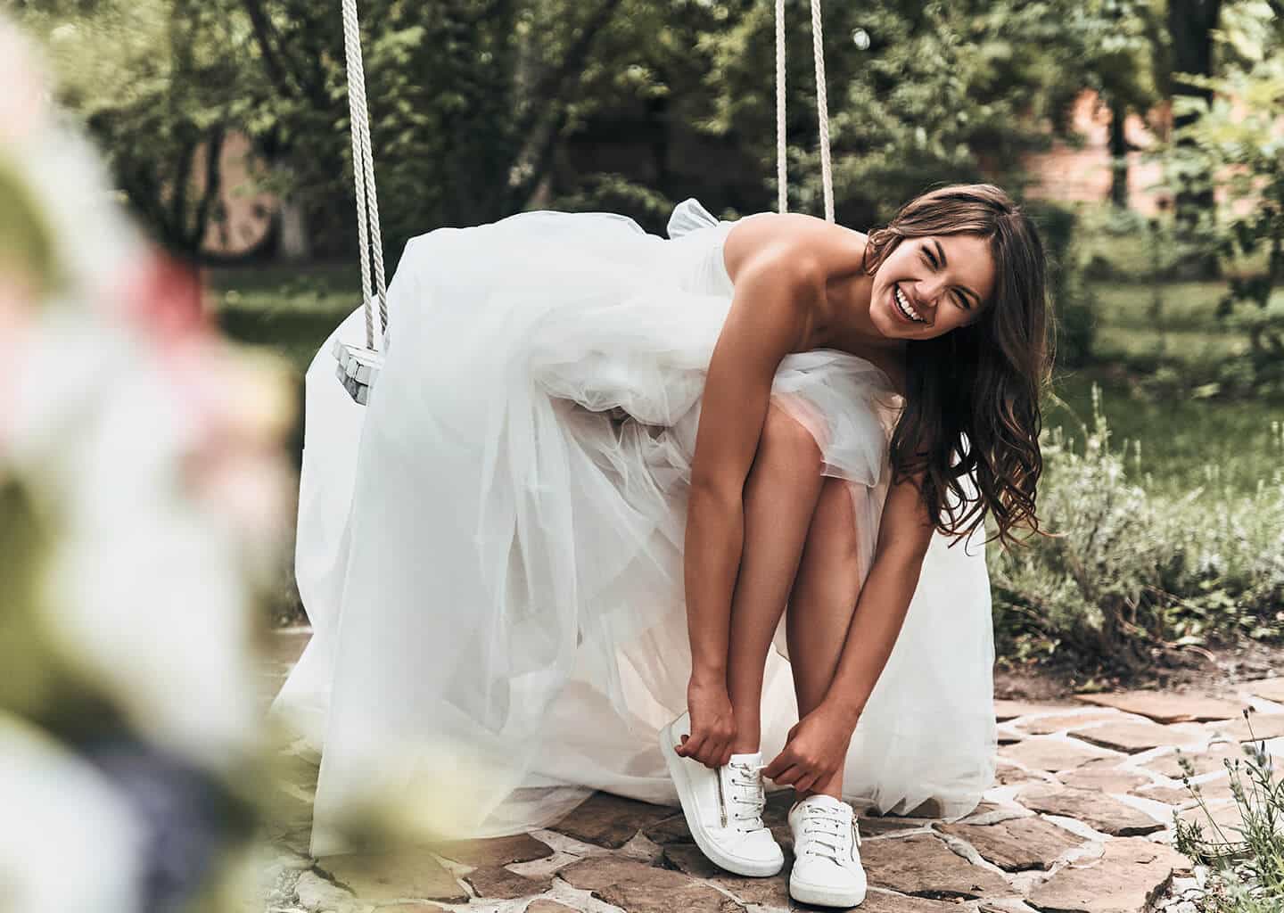 Gelin oturduğu salıncak üzerinde yere doğru eğilerek spor ayakkabılarının bağcıklarını bağlıyor ve gülümsüyor.
