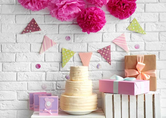 Açık renk arka plan önünde duran masa üzerinde üç katlı bir düğün pastası ve hediye kutuları bulunuyor. Duvarda ise pembe ve yeşil süsler duruyor.