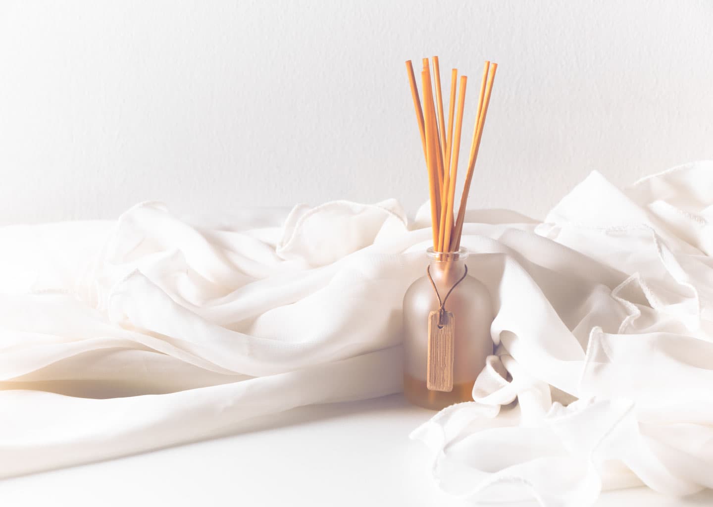 Açık renk plan üzerinde, beyaz bir tül ile sarılmış oda kokusu şişesi duruyor. Şişenin içerisinde tahta parçaları bulunuyor.