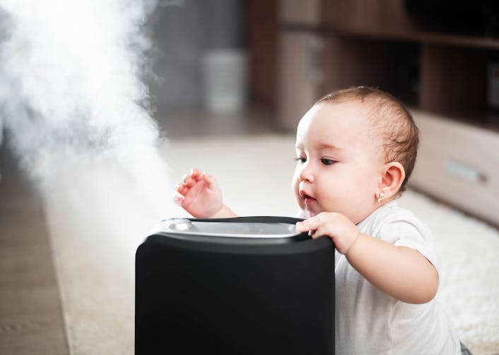 Yerde oturan bebek, önünde duran ve buhar üfleyen buhar makinesini tutuyor ve meraklı bir şekilde bakıyor.