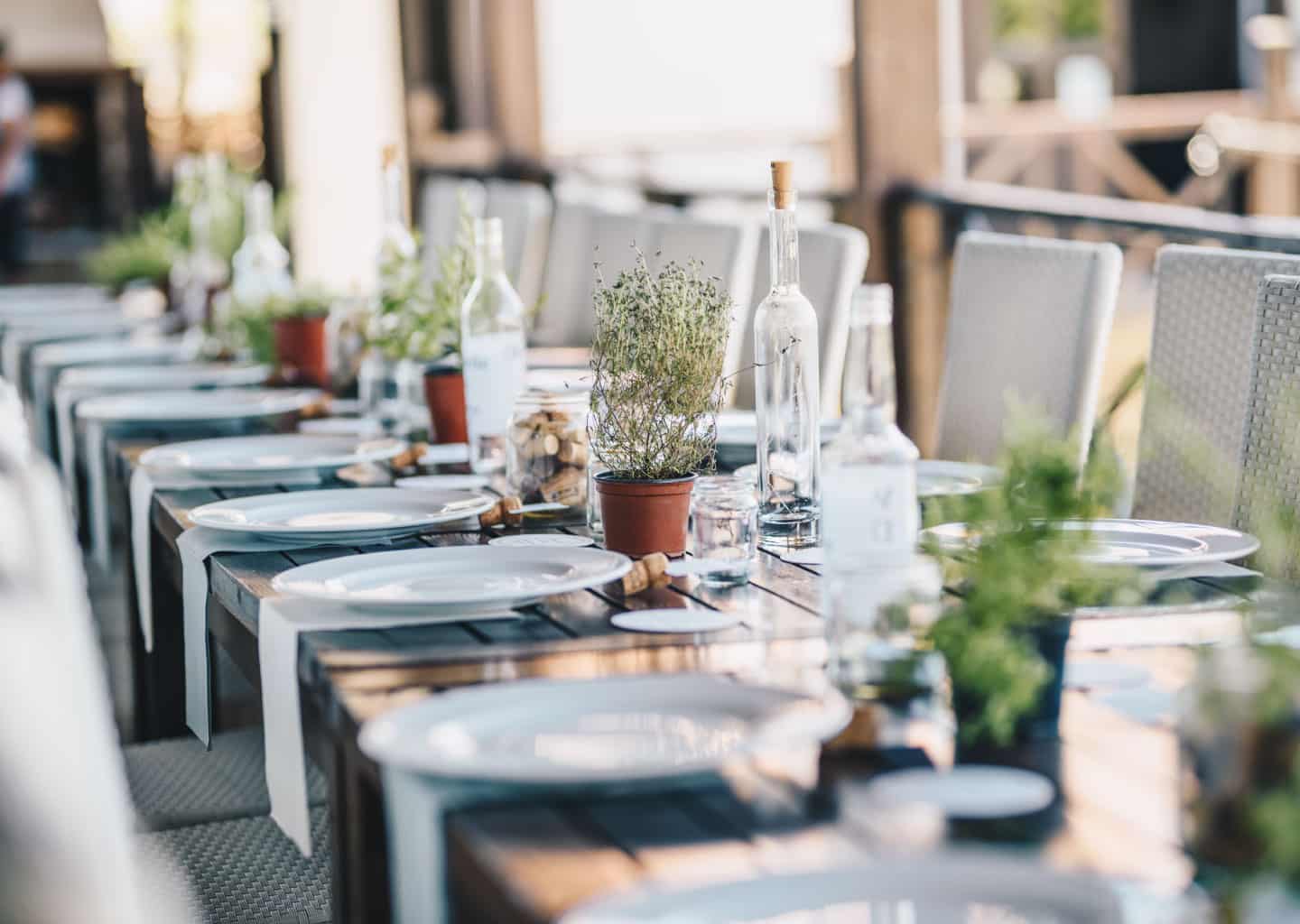 Yemek masası üzerinde; cam şişeler, tabaklar, tabak örtüleri ve dekoratif çiçekler bulunuyor. Masanın etrafında beyaz sandalyeler duruyor.