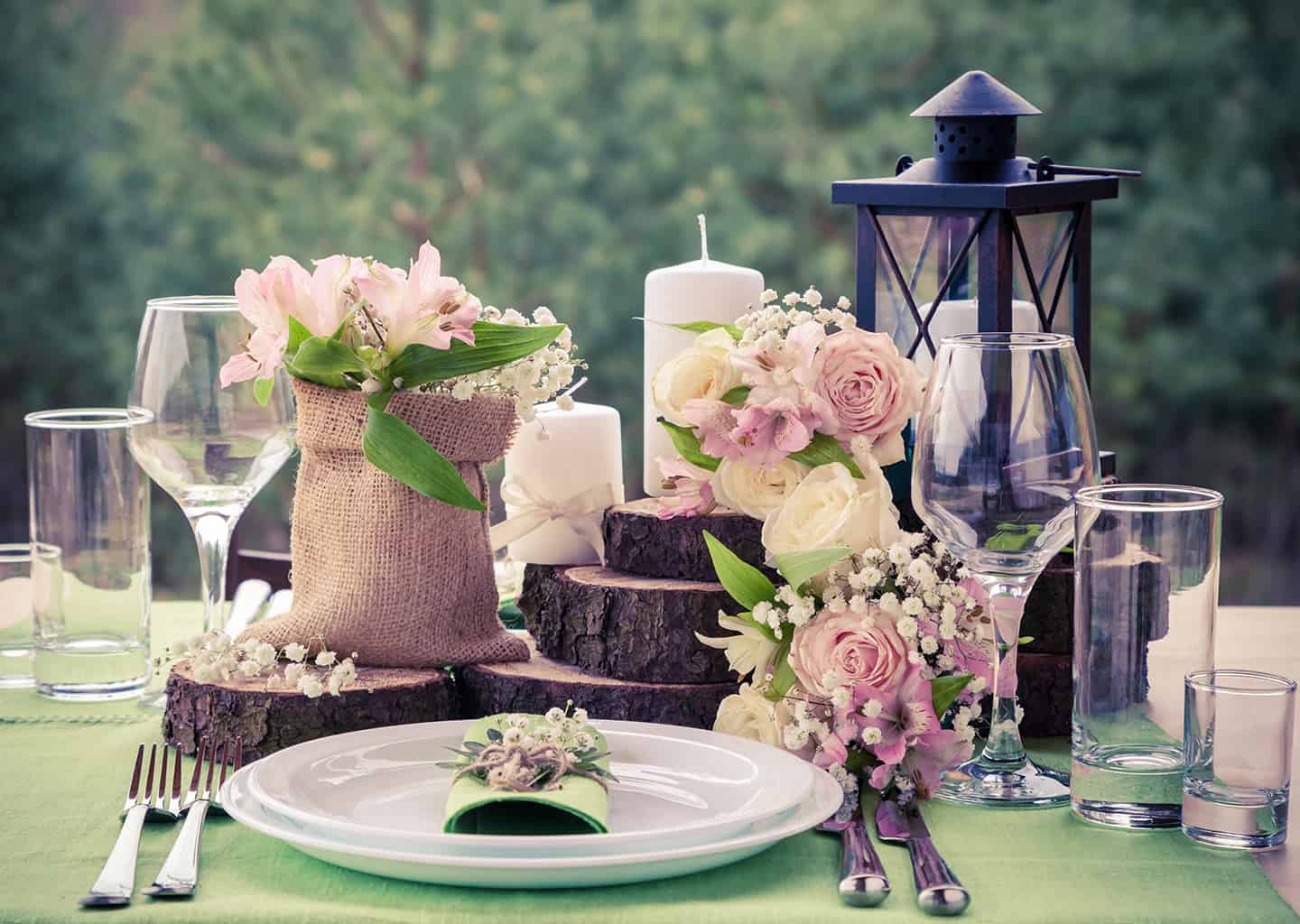 Yakın plan çekimde yemek masası üzerinde tabak, çatallar, bıçaklar, odundan yapılmış aksesuarlar ve çiçekler bulunuyor. Arka planda ormanlık alan görünüyor.