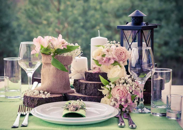 Yakın plan çekimde yemek masası üzerinde tabak, çatallar, bıçaklar, odundan yapılmış aksesuarlar ve çiçekler bulunuyor. Arka planda ormanlık alan görünüyor.