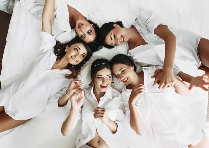 5 yakın kadın arkadaş, beyaz nevresimli bir yatak üzerinden dairesel olarak yatıyor. Kadınlar birbirlerine doğru bakarak gülümsüyor. Kadınların üzerinde beyaz gömlek bulunuyor.