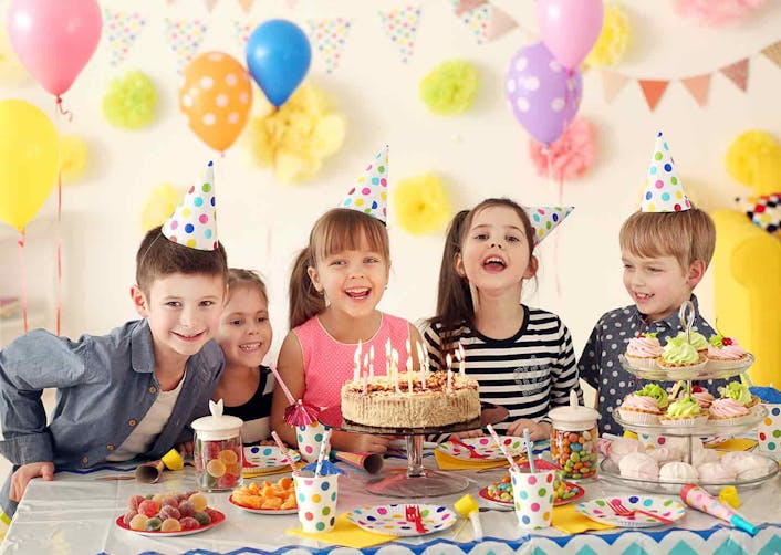 Çocuklar evde kafalarında bulunan doğum günü şapkaları ile doğum günü kutluyor. Duvarda renkli balonlar ve parti malzemeleri bulunuyor. Çocukların arkasında bulunduğu yemek masası üzerinde; mumlar ile çevrelenmiş bir pasta, karton bardaklar, minik kekler ve atıştırmalıklar bulunuyor.