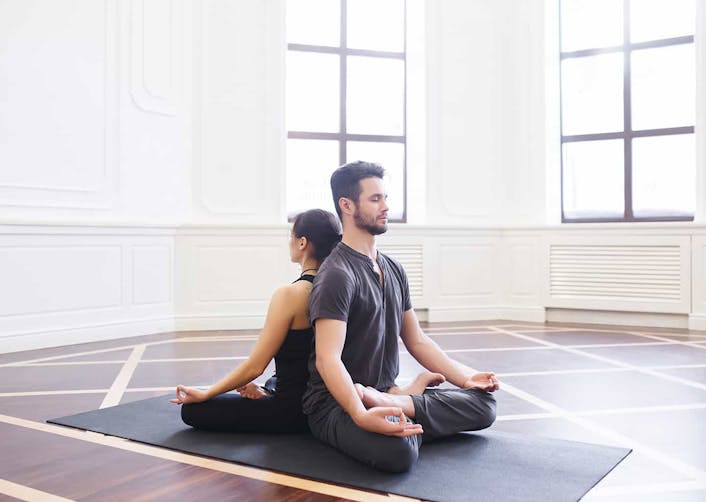 Kadın ve erkek etrafı boş olan bir salon içerisinde, yoga matı üzerinde sırt sırta vererek yoga yapıyorlar.