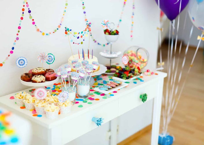Odada bulunan masa üzerinde parti için hazırlanmış olan rengarenk atıştırmalıklar bulunuyor. Masanın solunda ise yere tutturulmuş renkli balonlar duruyor.