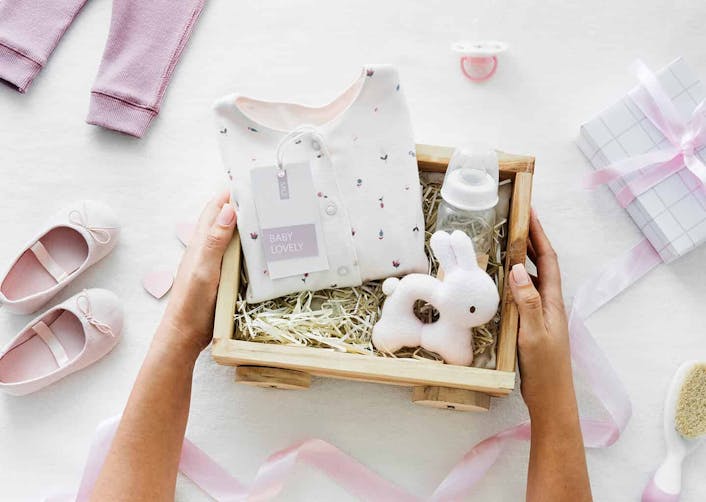 Yakın plan çekimde, beyaz arka plan üzerinde duran hediye kutusu içerisinde bebek giysisi, biberon ve oyuncak bir tavşan duruyor. Kutunun etrafında ise bebek ayakkabısı, bebek pantolonu ve pembe bir tül ile sarılmış hediye kutusu bulunuyor.
