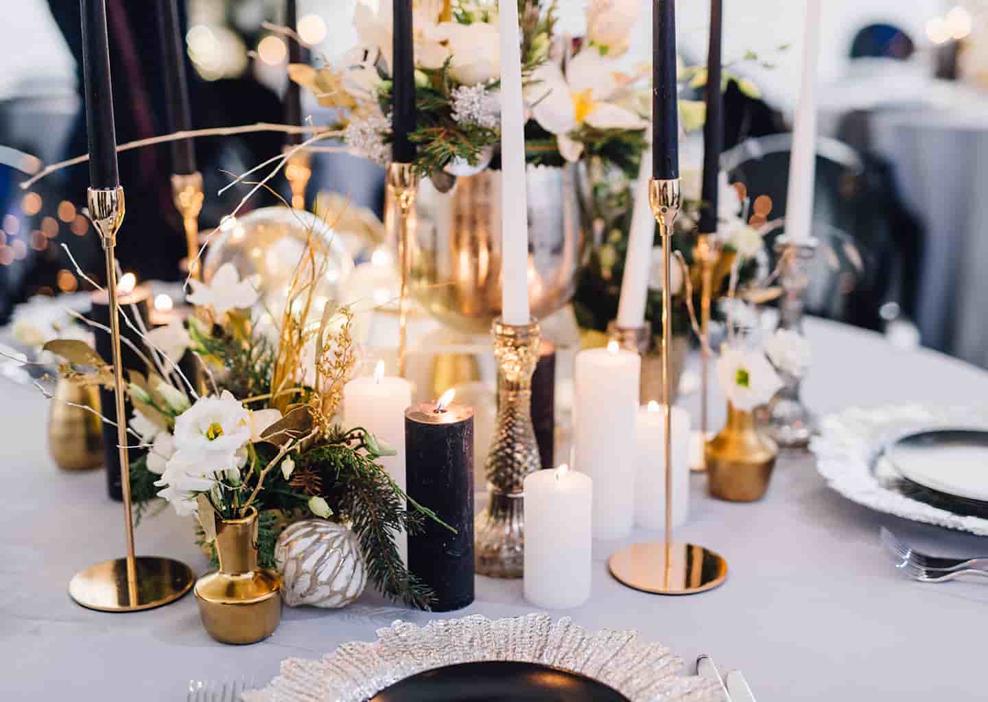 Yuvarlak yemek masası üzerinde mumlar, çiçekler, şamdanlar, tabaklar ve çatallar bulunuyor.