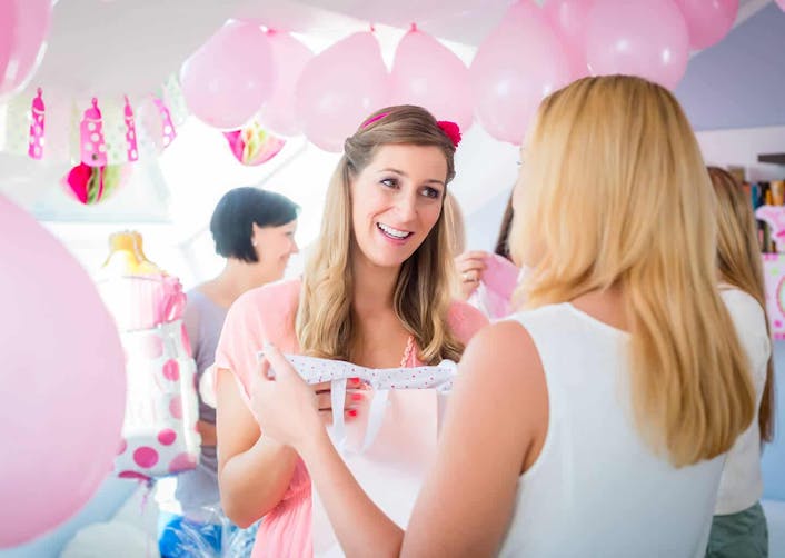 Baby shower partisi veren kadın, arkası dönük olan arkadaşı ile gülümseyerek sohbet ediyor. Davetlilerin bulunduğu odada pembe balonlar ve süslemeler bulunuyor.