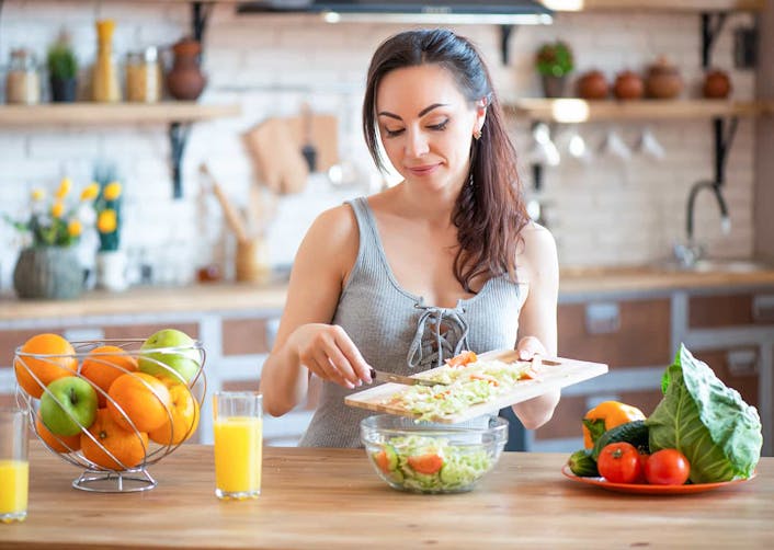 Mutfaktaki yemek masasında oturan genç kadın, kendisine salata hazırlıyor. Yemek masası üzerinde salata tabağı, şık bir tabak içerisinde elmalar ve portakallar, taze sıkılmış portakal suyu ve salata malzemeleri bulunuyor.