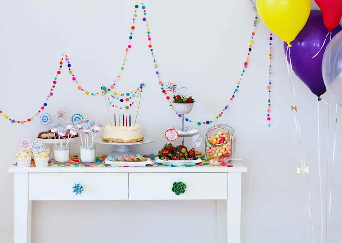 Açık renk arka plan önünde duran masa üzerinde, parti için için hazırlanmış renkli atıştırmalıklar ve pasta bulunuyor. Masanın solunda renkli balonlar duruyor.
