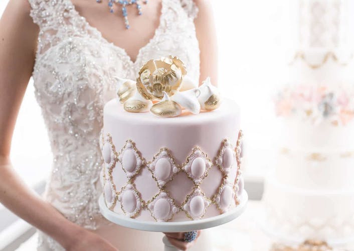 Yakın plan çekimde; askılı ve dantel detayları olan bir gelinlik giyen gelinin önünde düğün pastası bulunuyor.
