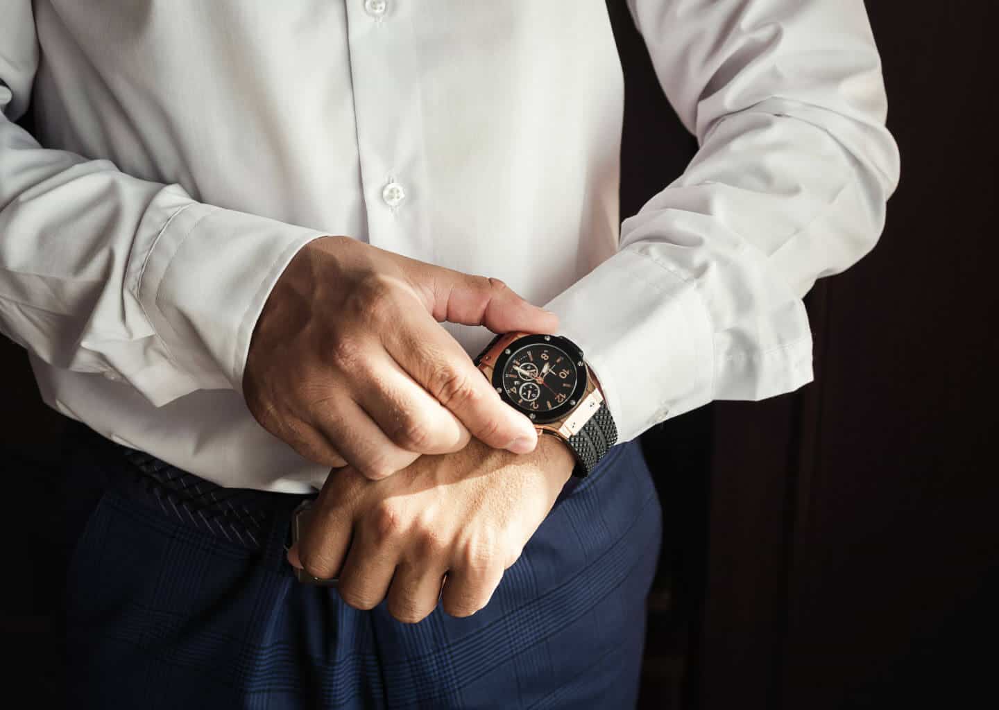 Yakın plan çekimde damat sol bileğinde bulunan saatini sağ eli ile tutuyor. Damadın üzerinde beyaz bir gömlek ve lacivert bir pantolon bulunuyor.