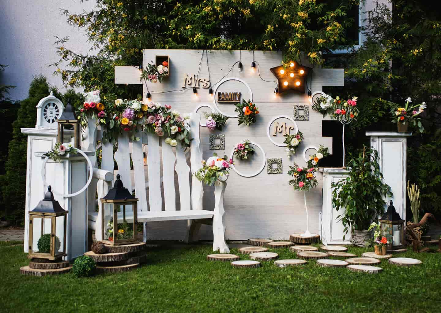 Bahçede beyaz renge boyanmış tahtalardan oluşturulan bir duvar, çiçekler, ışıklandırmalar ve aksesuarlar bulunuyor.