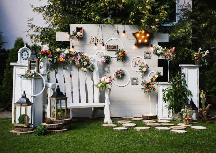 Bahçede beyaz renge boyanmış tahtalardan oluşturulan bir duvar, çiçekler, ışıklandırmalar ve aksesuarlar bulunuyor.