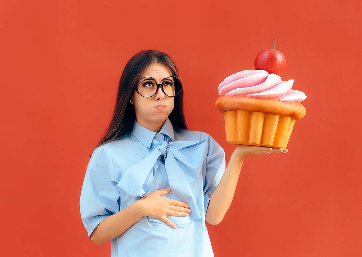 Koyu renk arka plan önünde duran genç kadın elinde oyuncak bir pasta tutuyor. Kadın bir eli ile pastayı tutarken diğer eli ile de midesini tutuyor. Kadının üzerinde mavi ve kısa kollu bir gömlek bulunuyor.