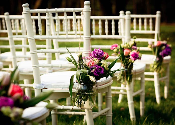 Yakın plan çekimde; bahçe içerisinde düğün töreni için yerleştirilmiş olan beyaz sandalyeler bulunuyor. Sandalyelerin sağ bölümünde çiçek buketleri bulunuyor.
