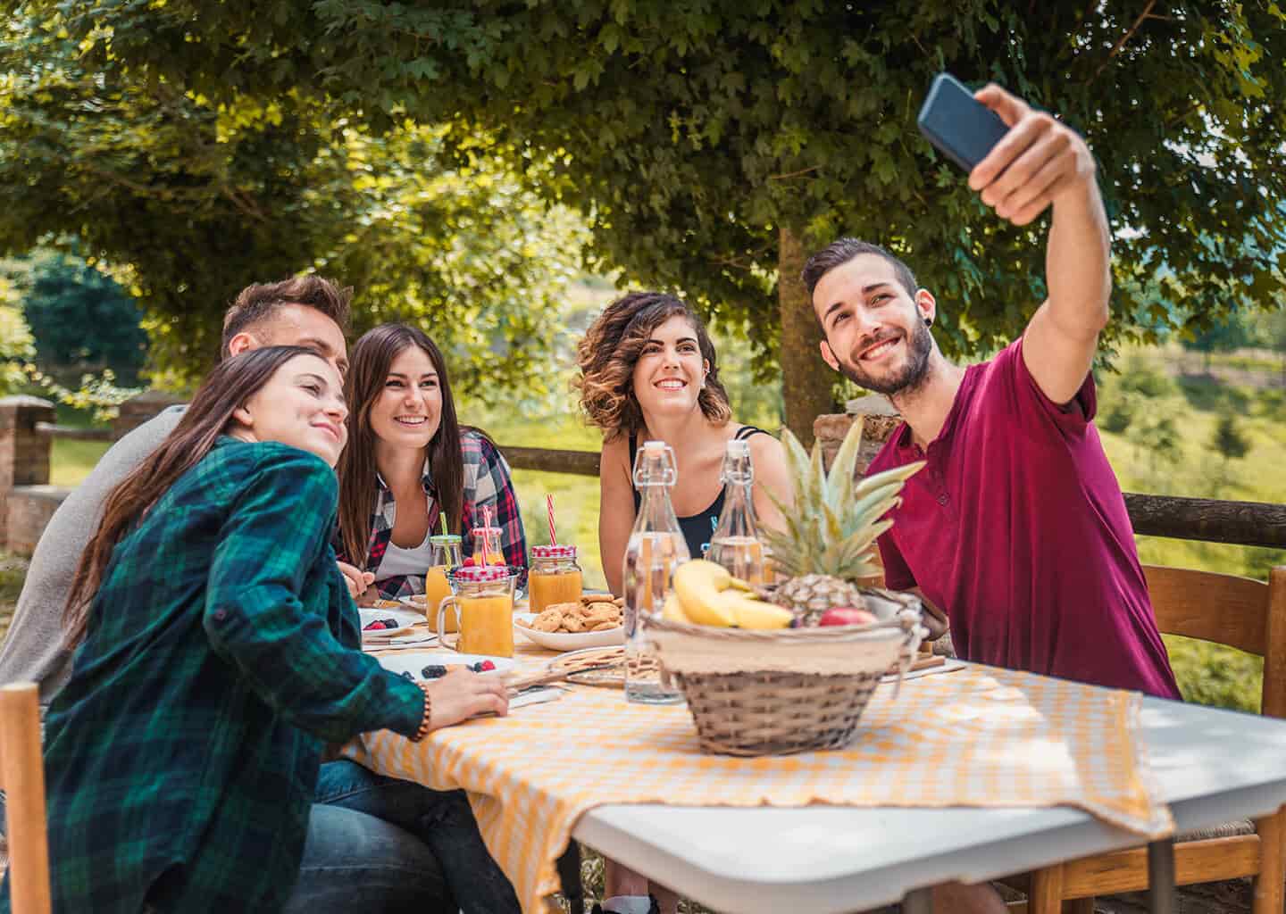 Ormanlık bir alan içerisinde, yemek masası etrafında toplanmış ve yemek yiyen arkadaş grubundan bir kişi, arkadaşları ile fotoğraf çekiyor.