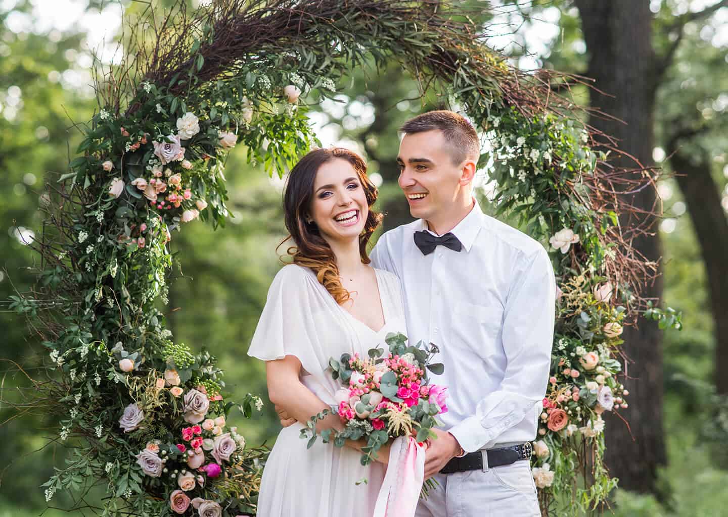 Gelin ve damat birbirine sarılıyor. Gelin ve damadın arkasında yuvarlak ve yeşilliklerden yapılmış bir aksesuar bulunuyor.