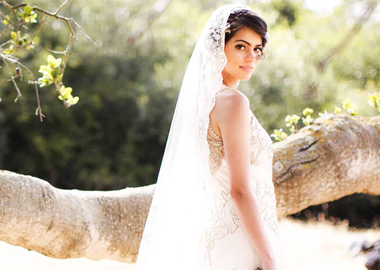 Gelin ormanlık bir alanda, bir ağaç dalının önünde yan dururken sağına doğru bakarak gülümsüyor, gelinin kafasında uzun bir duvak ve üzerinde dantelli bir gelinlik bulunuyor.