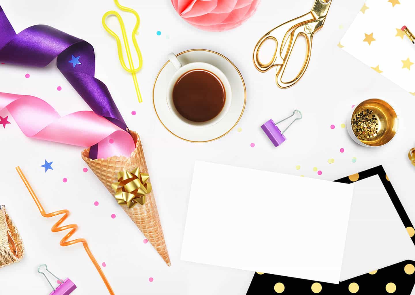 Beyaz renk arka plan üzerinde bir fincan kahve, makas, kağıt, parti malzemeleri ve davetiye zarfı bulunuyor.