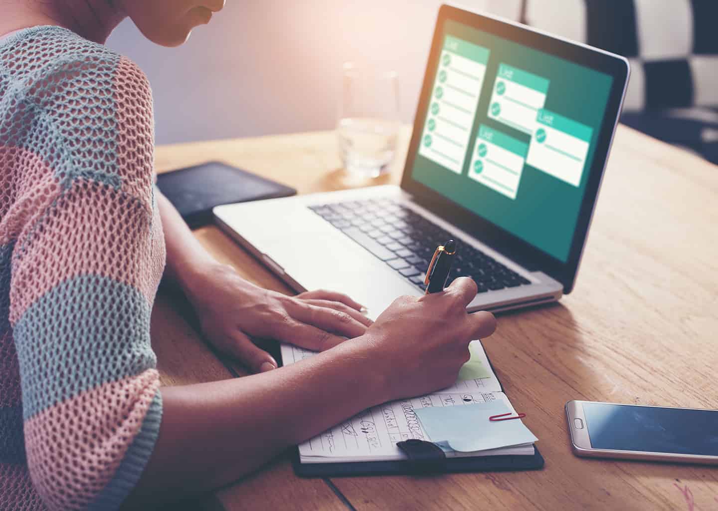 Yakın plan çekimde kadın; ahşap bir masa üzerinde duran laptopa bakarak not defterine notlar alıyor. Masa üzerinde bir tablet, akıllı telefon ve su bardağı bulunuyor.