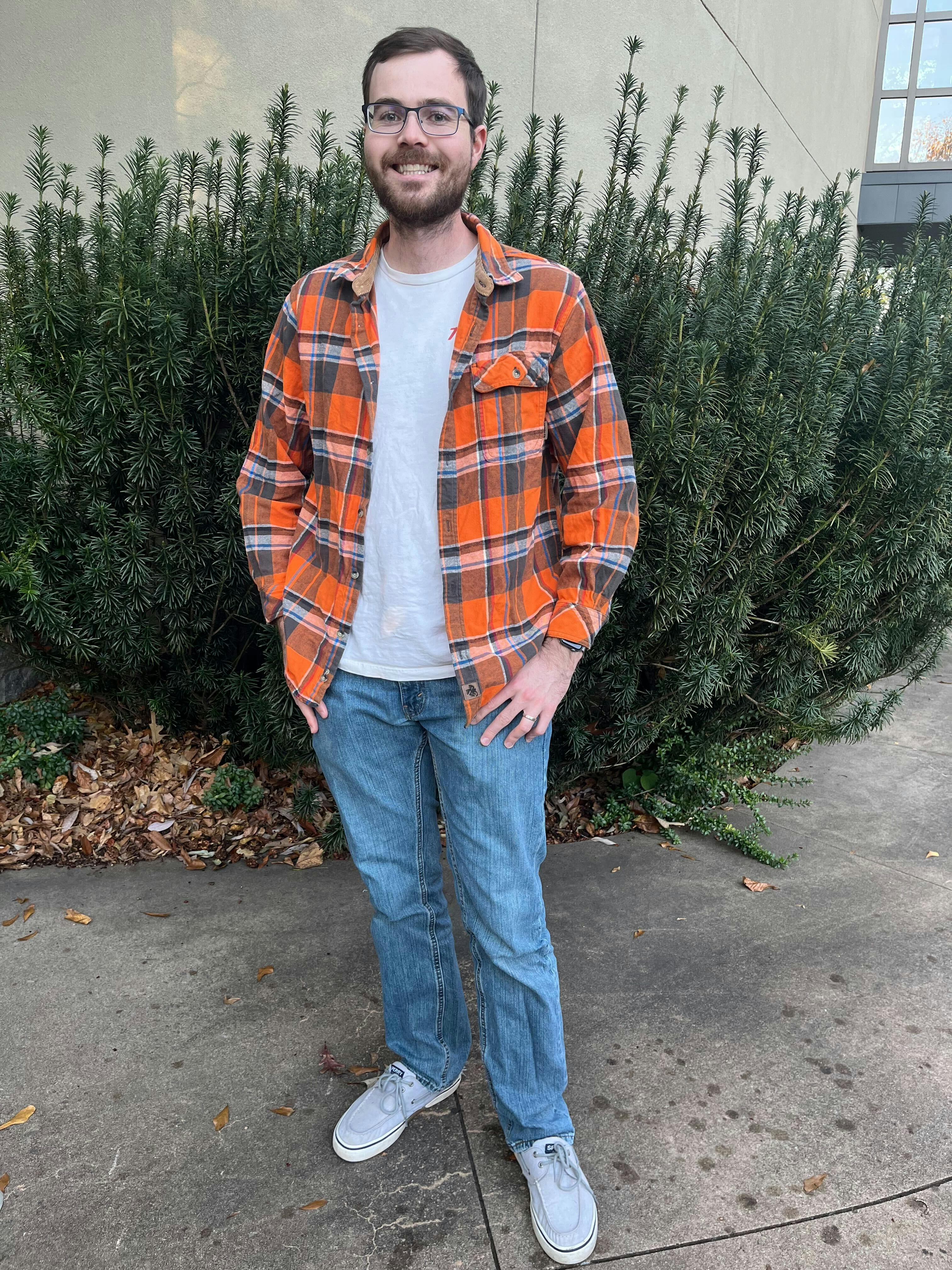 The man puts his hands into his pockets and stands by a big green bush