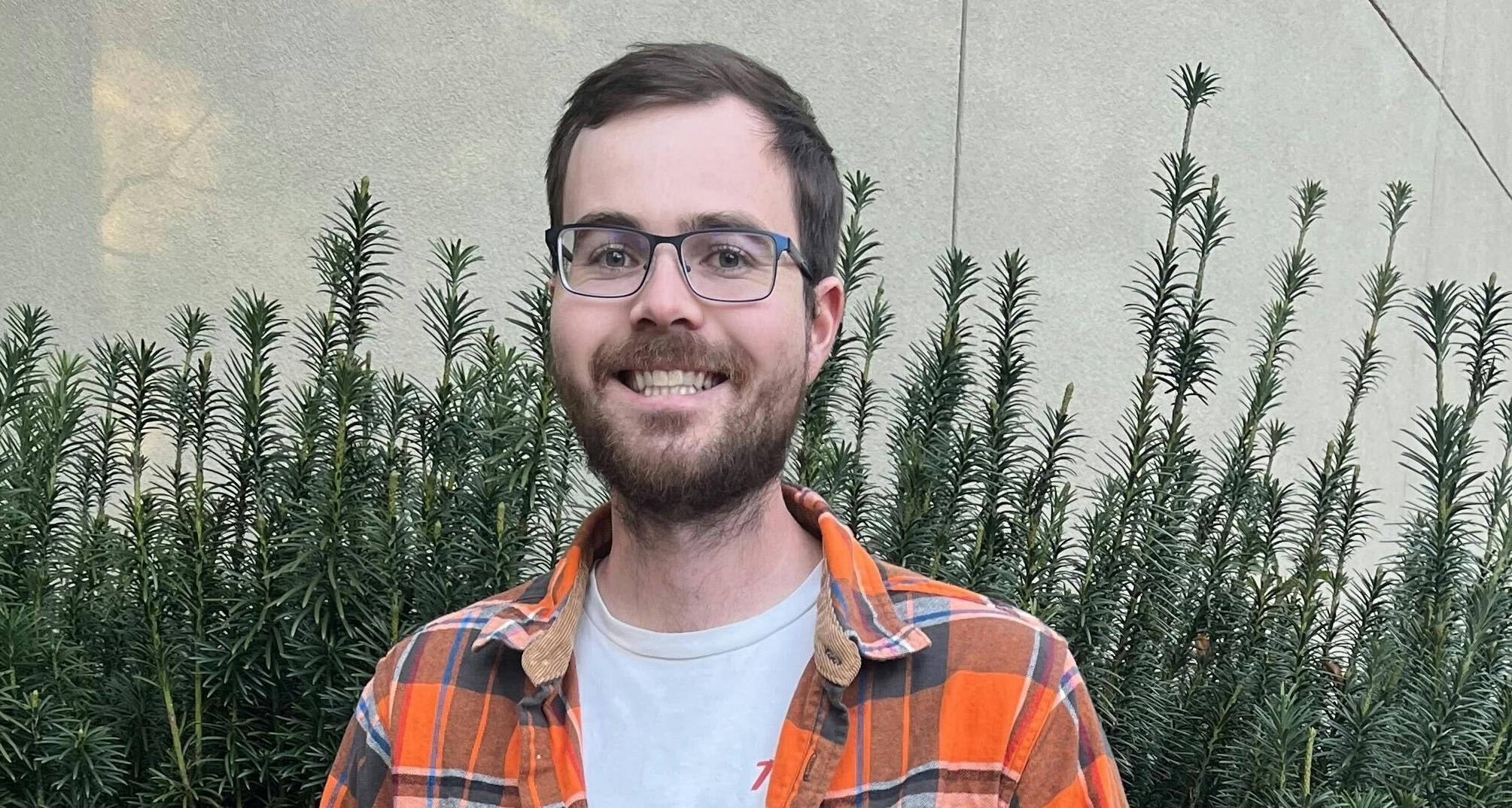Photo of Matt with glasses, brown hair and a beard. He's wearing an orange plaid shirt over a white t-shirt and smiling. There is a plant behind him. 